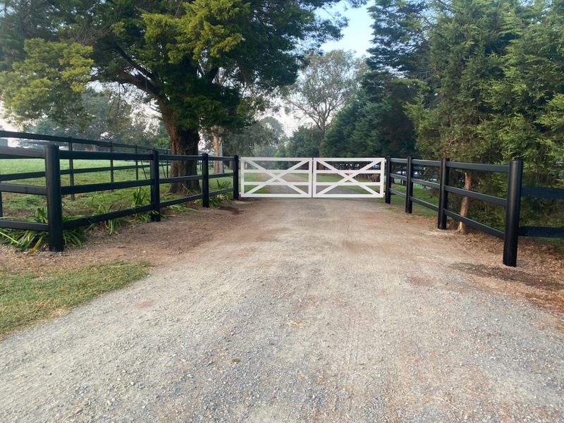 White steel farm entrance gates on a gravel driveway with black post-and-rail fencing